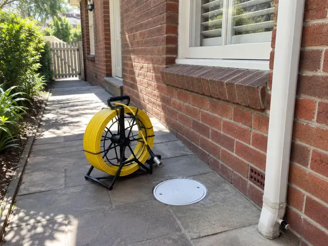 Capped drain inspection opening beside a yellow camera reel on a paved brick house side path in Killara