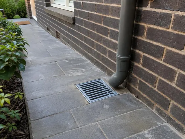Outdoor gully grate beside a brick house wall with paved path in Killara